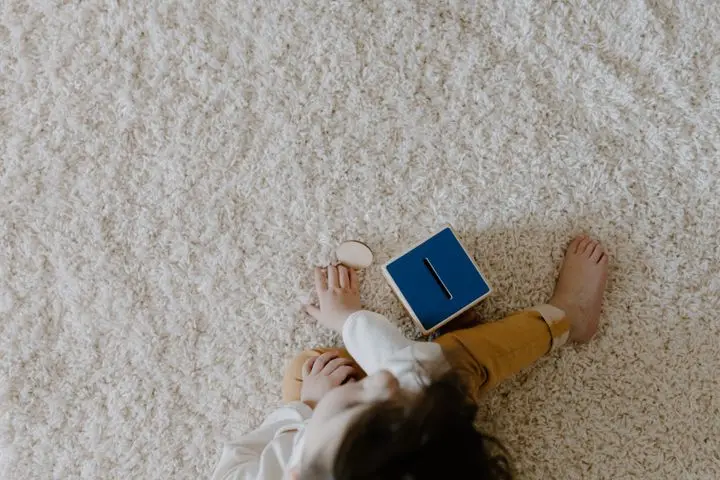 A young child sits on a carpet, engaging with wooden toys and a blue box, captured in a cozy indoor setting.