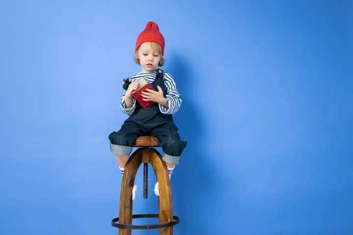 Cute child in a red hat reading on a wooden stool against a blue background.