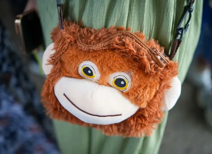 Adorable monkey face plush bag worn by a person in green, offering a fun fashion accessory.