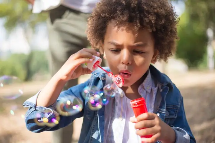 A young boy blowing bubbles outside in a sunny park, enjoying a playful moment.