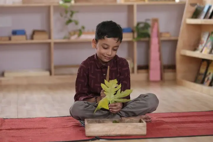 A young boy sitting on a mat examining a leaf in a classroom environment.