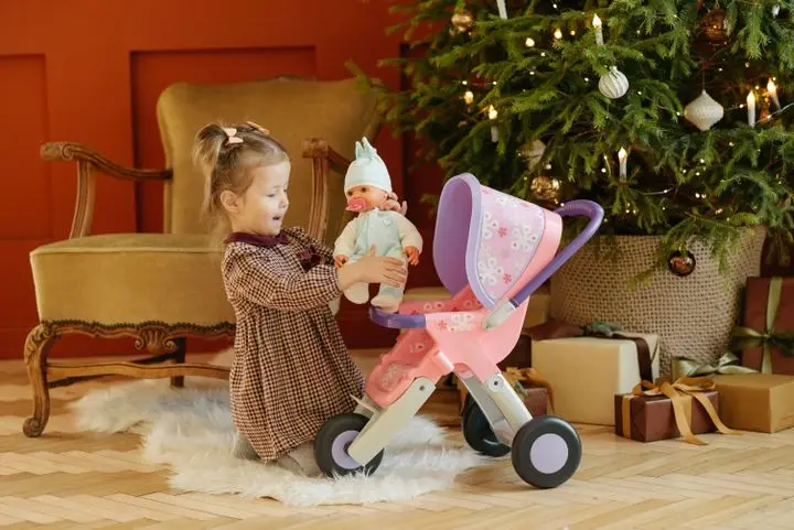 Adorable child playing with a doll stroller next to a decorated Christmas tree indoors.