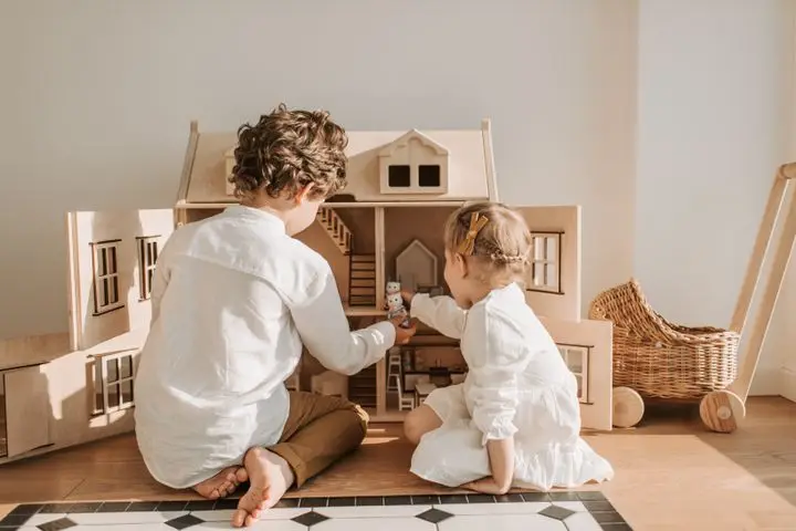 Two children enjoy playing with a wooden dollhouse in a sunlit room, showcasing sibling bonding.