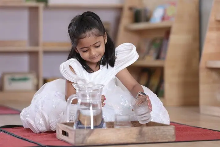 A young girl exploring Montessori activities with a tray indoors, dressed in a white dress.