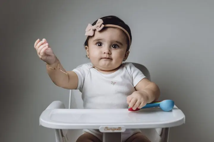 Cute baby girl in a high chair with a bow hairband, extending her hand playfully in a studio setting.