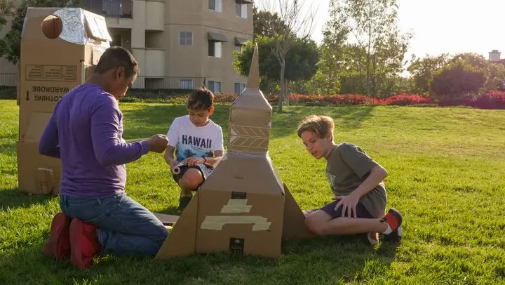 Three boys enjoying a sunny day building a cardboard rocket in a park, showcasing creativity and teamwork.