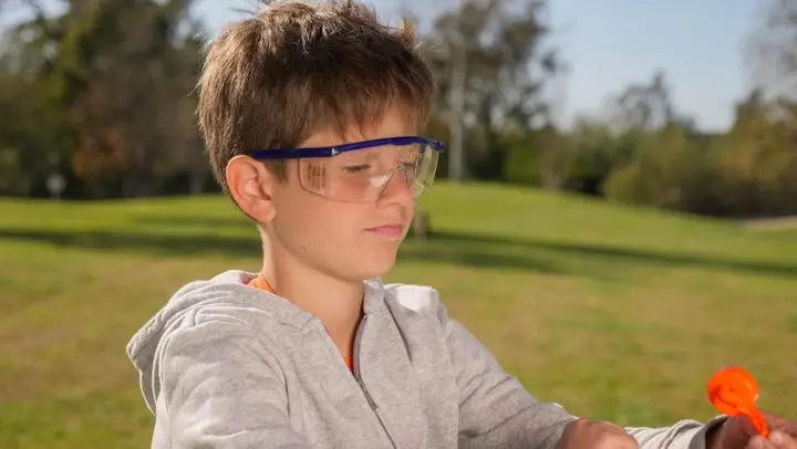 A young boy wearing a gray sweater and protective goggles playing with a toy outside on a sunny day.