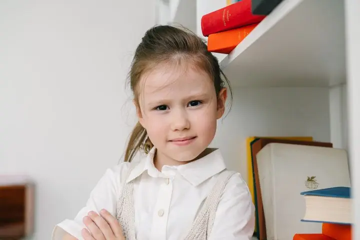 Cute young girl confidently posing with arms crossed next to a bookshelf.