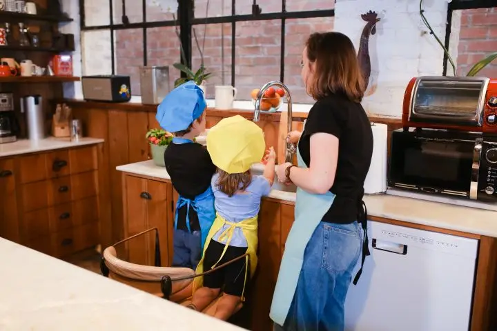 Mother and children washing hands in a cozy kitchen, wearing colorful chef hats.