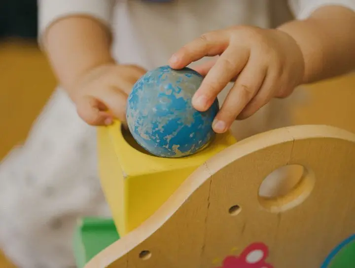 Close-up of child's hands placing a blue ball on a yellow wooden toy, emphasizing play and development.