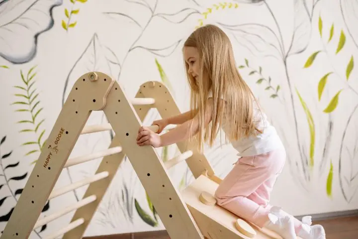 A young girl with long hair climbing an indoor wooden play ladder in a cozy room.