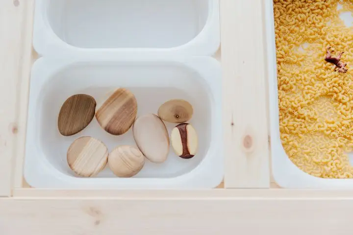 Set of various white plastic containers with stones and raw pasta for child sensory development on wooden table in light room