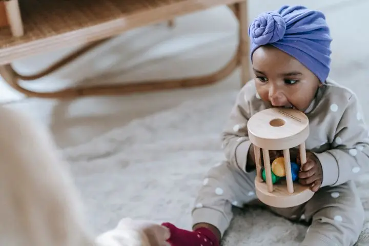 Adorable African American child in headwear with wooden toy sitting on floor near crop parent in light room at home on blurred background