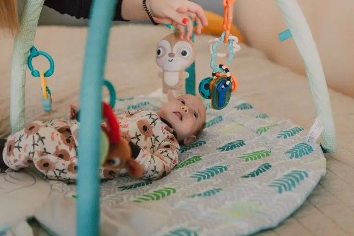 Cute baby lying on a play mat wearing a onesie and interacting with hanging toys.