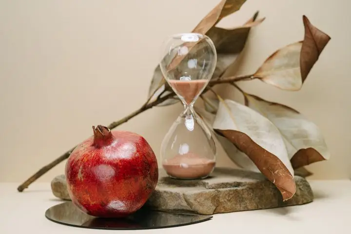 A pomegranate and hourglass on a stone surface with autumn leaves, symbolizing time and nature.