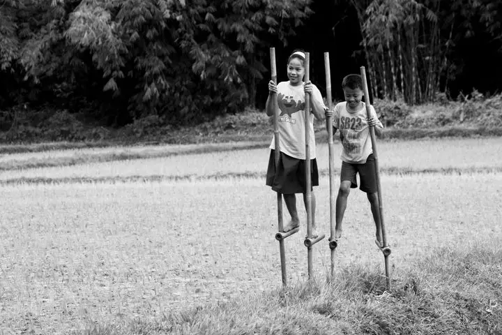Black and white photo of children playing on stilts in a rural setting, joyful and carefree.
