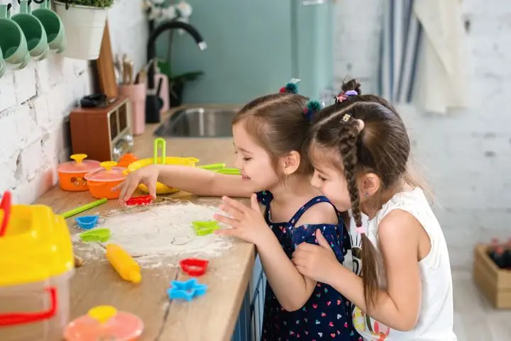 Two girls playing and baking with flour and colorful tools in a kitchen setting.
