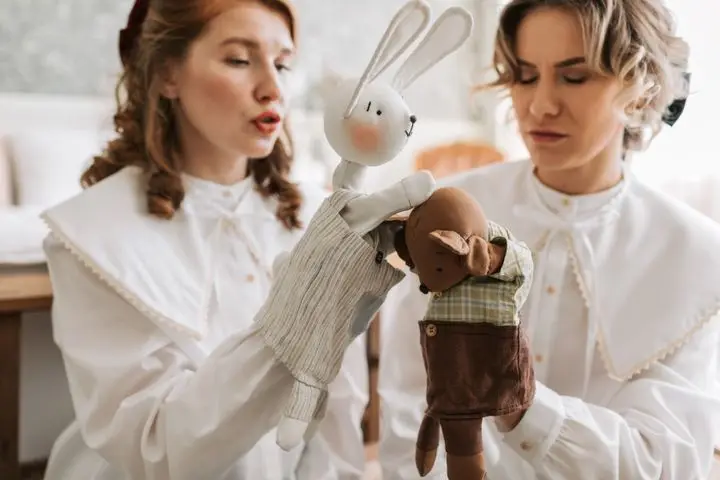 Two women in white attire engaging in an indoor puppet show with plush toys.
