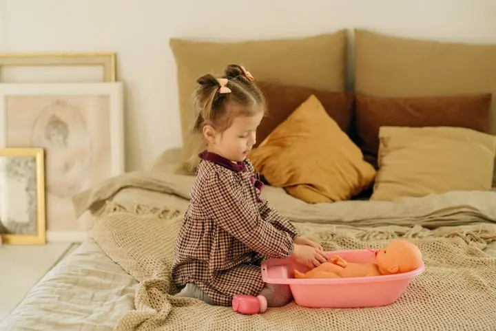 Little girl sitting on bed playing with a doll in a cozy indoor setting, highlighting childhood leisure.
